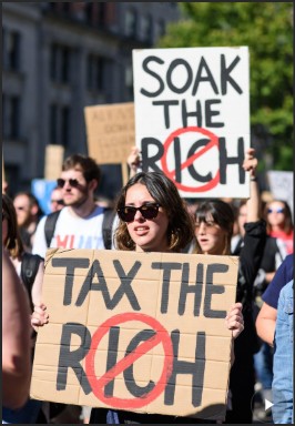 A protestor holds a sign that reads 'TAX THE RICH,' with other attendees in the background, showcasing a public demonstration advocating for higher taxes on wealthy individuals.