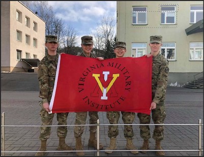 Four military personnel in uniform holding a red flag with the Virginia Military Institute emblem in an outdoor setting.