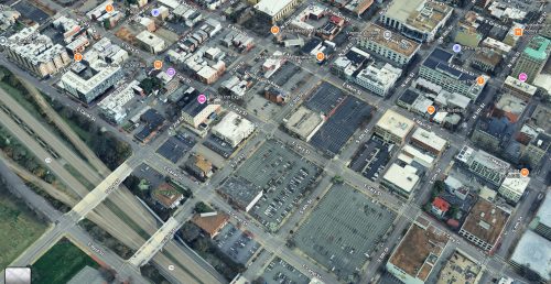 Aerial view of a city block, showing a grid layout with multiple buildings, streets, and parking areas nearby. Several roads and highways are visible, along with various commercial establishments.