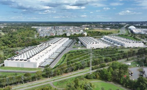 Aerial view of a large industrial facility with multiple buildings and rooftop cooling units, surrounded by greenery and power lines.