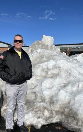 A man wearing sunglasses and a black jacket stands next to a large pile of snow in a parking lot, with a blue sky in the background.
