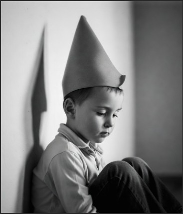 A black and white image of a young boy wearing a cone-shaped hat, sitting against a wall with a thoughtful expression.