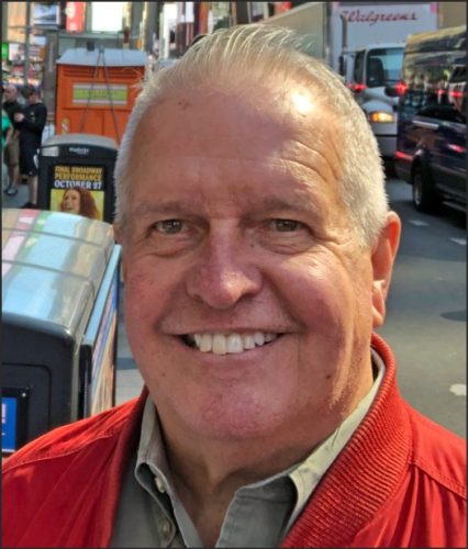 A smiling older man with gray hair wearing a red jacket, standing on a busy street in an urban setting.