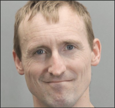 Close-up portrait of a smiling man with short blonde hair and blue eyes against a neutral background.