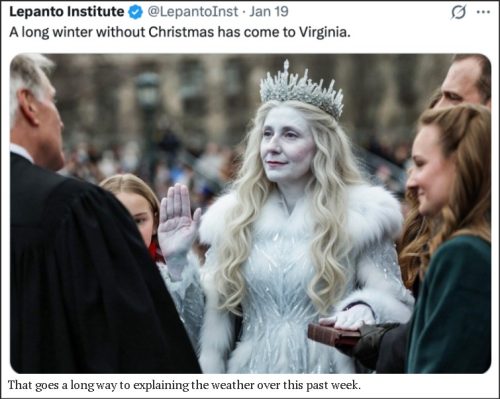 A woman dressed as a winter queen with long white hair, a sparkling crown, and a white fur coat stands in front of an audience, raising her right hand. She is surrounded by other people in formal attire, with a focus on her regal appearance.
