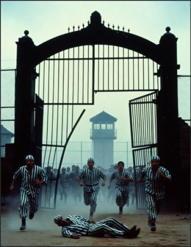 Group of prisoners in striped uniforms running through a large open gate, with a guard tower in the background and a figure lying on the ground.