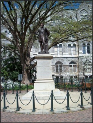 A bronze statue of a historical figure on a stone pedestal, surrounded by a circular chain fence and large trees, with a historic building in the background.