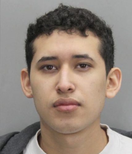 A close-up mugshot of a young man with curly black hair, wearing a gray hoodie and a white shirt, looking directly at the camera.