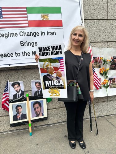 A woman holds a sign reading 'Make Iran Great Again' at a protest, standing in front of a display featuring American flags and photos of individuals, with a backdrop stating 'Regime Change in Iran Brings Peace to the Middle East.'