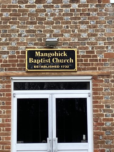 Sign for Mangohick Baptist Church established in 1732 on a brick wall above double doors.