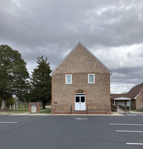 A brick building with a peaked roof and white windows, set against a cloudy sky, surrounded by trees and a parking lot.
