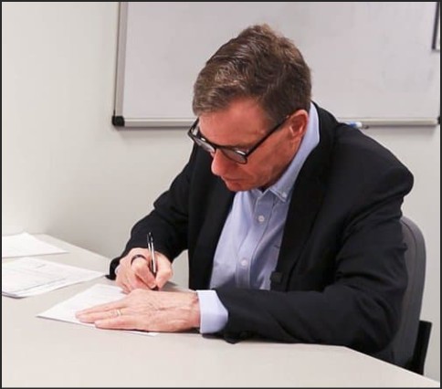 A man in a black suit and glasses is sitting at a table, writing on a piece of paper.