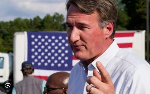 A man gestures while speaking outdoors, with a large American flag displayed in the background.