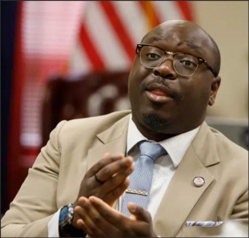 A man in a light gray suit and glasses gestures while speaking, with an American flag blurred in the background.