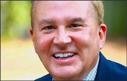 Close-up portrait of a smiling man with short light-colored hair, wearing a dark blazer and a checked shirt.