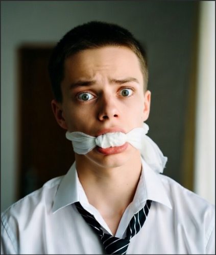 A young man with short hair and a surprised expression, wearing a white shirt and a tie, has his mouth gagged with a piece of cloth.