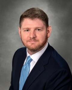 A professional portrait of a man with a short beard, wearing a dark suit and a light blue tie, against a soft gray background.