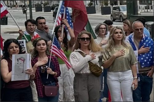 A diverse group of people standing together outdoors, some holding flags, with their hands over their hearts, expressing solidarity and unity.