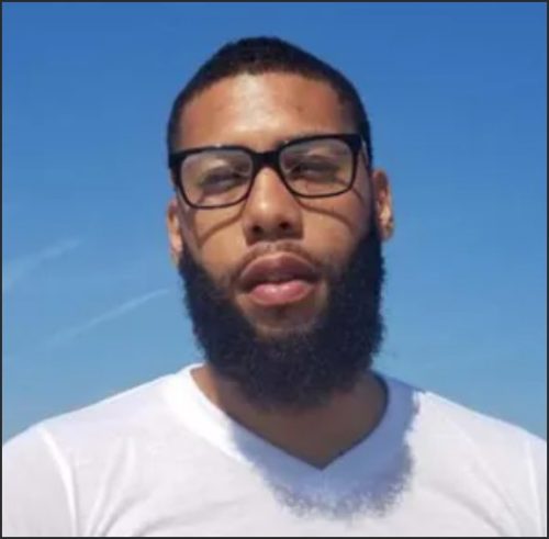 A man with a beard and glasses standing outdoors against a clear blue sky.