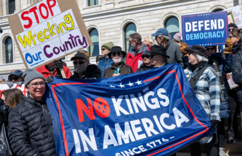 A group of protesters holding signs at a demonstration, with one sign reading 'STOP Wrecking Our Country' and another stating 'Defend Democracy'. A large banner reads 'NO KINGS IN AMERICA' with a historical reference to 1776.
