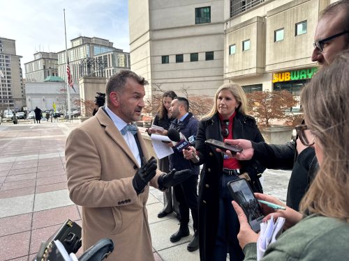 A man in a light coat speaks to reporters outside a courthouse, surrounded by journalists with microphones and notepads. The scene captures a busy atmosphere with urban buildings in the background.