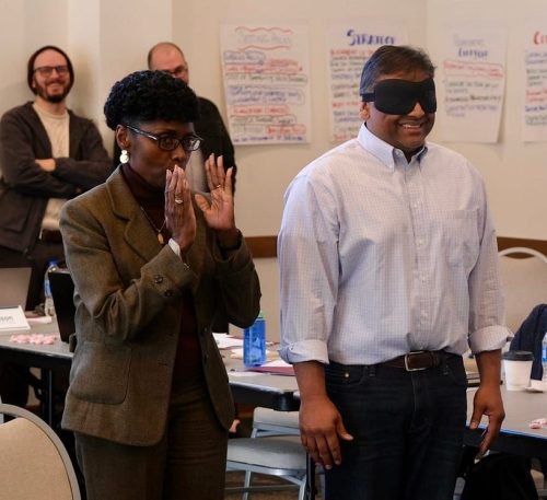 A woman in a suit gestures with her hands while standing next to a blindfolded man wearing a collared shirt, both participating in a group activity in a meeting room.