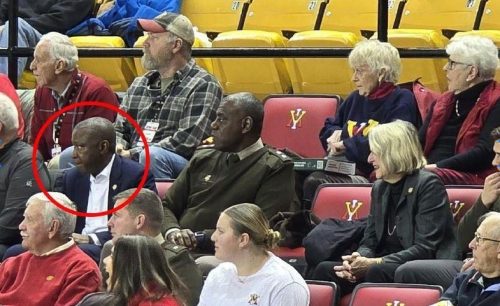 A group of spectators sitting in bleachers at an event, with a focus on a man in a white shirt and black jacket, seated among others.