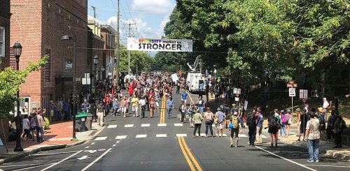 A street filled with a large crowd of people participating in a march, with buildings on either side and a banner above reading 'Diversity Makes Us Stronger'.