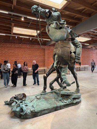 A large bronze sculpture depicting a rearing horse and its rider in an art gallery, with visitors observing the artwork in the background.