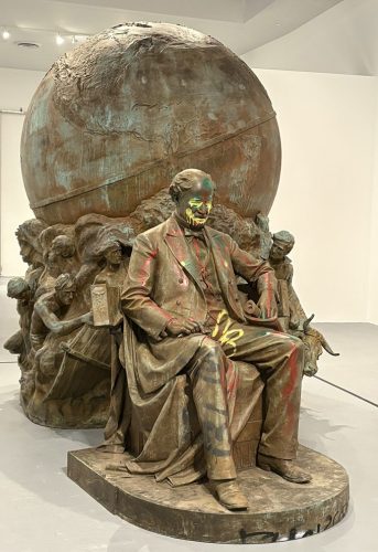 A sculpture depicting a seated man in formal attire, surrounded by smaller figures, with a large globe behind him. The statue is covered in colorful paint, giving it a vibrant, modern appearance.