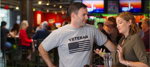 A man wearing a grey 'VETERAN' t-shirt with an American flag stands casually talking to a woman in a bar setting, with other patrons visible in the background.
