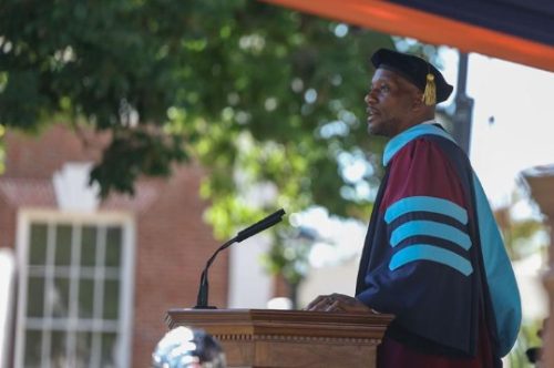 A speaker in academic regalia giving a speech at a graduation ceremony, standing behind a wooden podium with a microphone, surrounded by greenery.