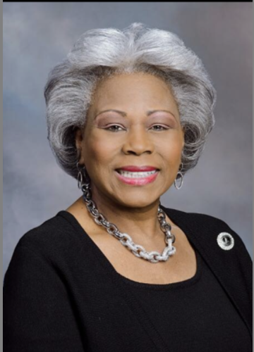 A professional portrait of an older woman with gray hair styled in a wavy fashion, wearing a black top and a large silver chain necklace. She is smiling and facing the camera.