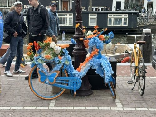 A blue bicycle decorated with colorful artificial flowers, positioned near a canal with a lamppost and people walking by.