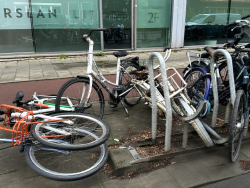 A mixed cluster of bicycles leaning against a bike rack, with some bikes appearing abandoned or broken, located on a city sidewalk.