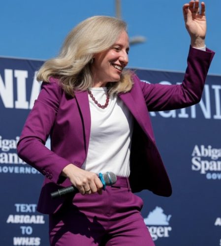 A woman in a purple suit joyfully raising her hand while speaking into a microphone at an outdoor event.