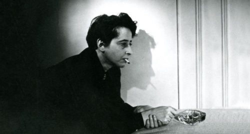 A black and white image of a person sitting against a wall, with a cigarette in their mouth and a pensive expression. They are holding a glass ashtray with their right hand.