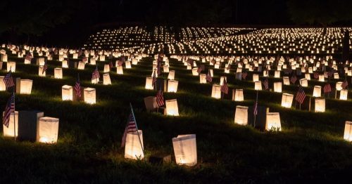 A serene nighttime scene of a cemetery illuminated by numerous glowing luminary bags, each marked with a small American flag, creating a poignant tribute.