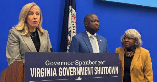 Three individuals at a podium discussing the Virginia Government Shutdown, with a blue background and a Virginia state flag in view.