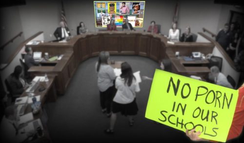 A group of people at a public meeting, with two individuals in the foreground holding a sign that reads 'NO PORN IN OUR SCHOOLS'. In the background, a screen displays various images, and the meeting participants are seated in a semicircle.
