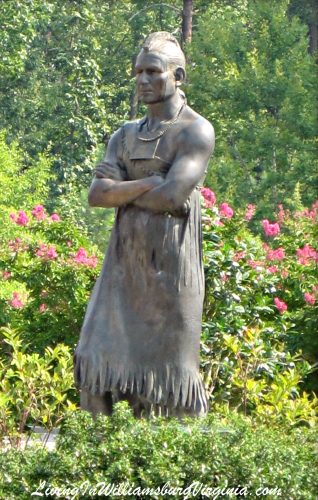 A bronze statue of a Native American man standing with arms crossed, wearing a traditional outfit with fringed edges, surrounded by green foliage and flowering bushes.