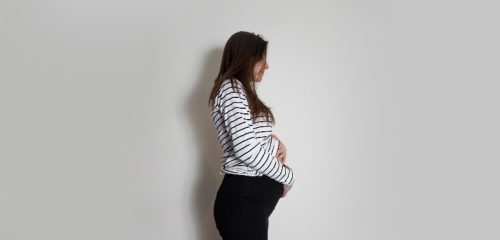 A pregnant woman in a striped long-sleeve shirt and black pants gently cradles her baby bump while standing against a plain background.