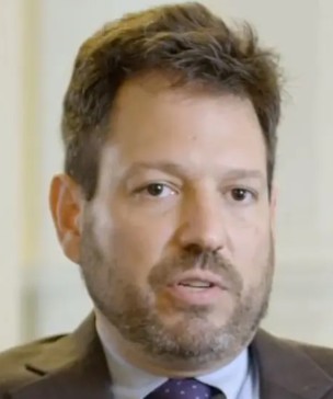 Close-up of a man with a beard, wearing a brown suit jacket and a blue shirt, speaking or engaged in conversation.