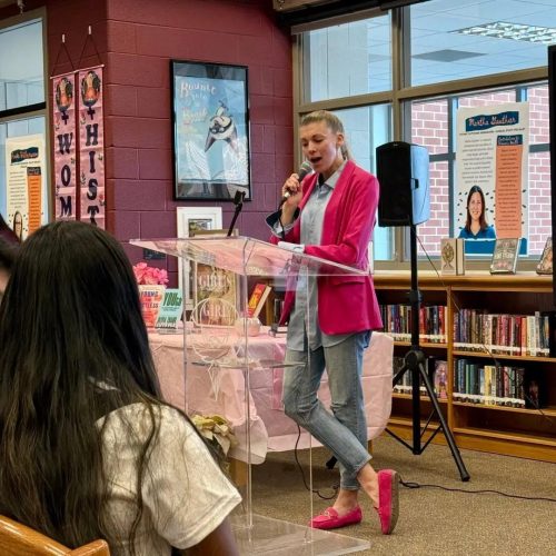 A young woman in a bright pink blazer speaking into a microphone at a podium in a library setting, with shelves of books and promotional posters in the background.