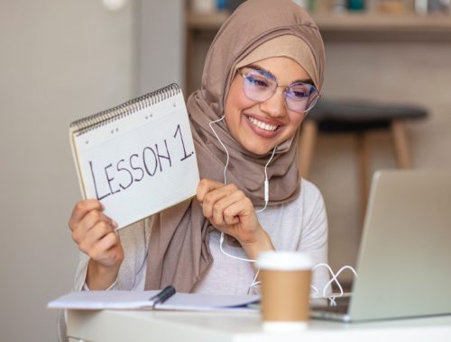 A smiling woman wearing a hijab and glasses holds up a notebook with 'LESSON 1' written on it, while sitting at a desk with a laptop and a coffee cup.