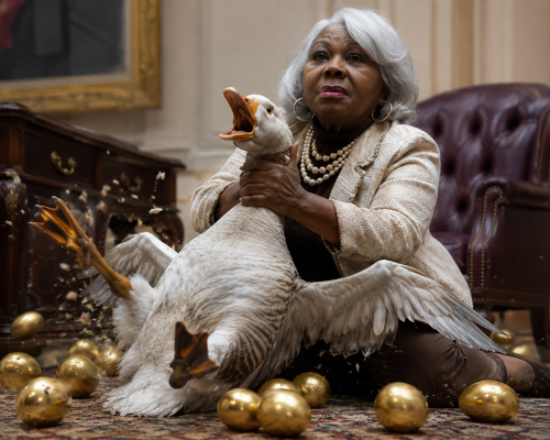 An elderly woman with silver hair is holding a large goose while sitting on a patterned rug. Surrounding her are several golden eggs scattered across the floor.