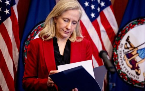 A woman in a red blazer is reading a document while standing in front of Virginia state flags.